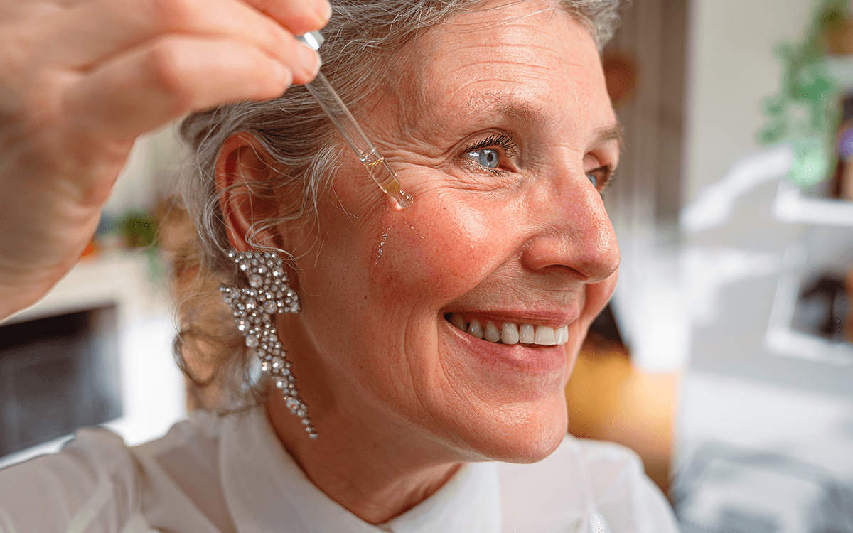 En äldre kvinna med vitt hår som applicerar serum i ansiktet med hjälp av en pipett